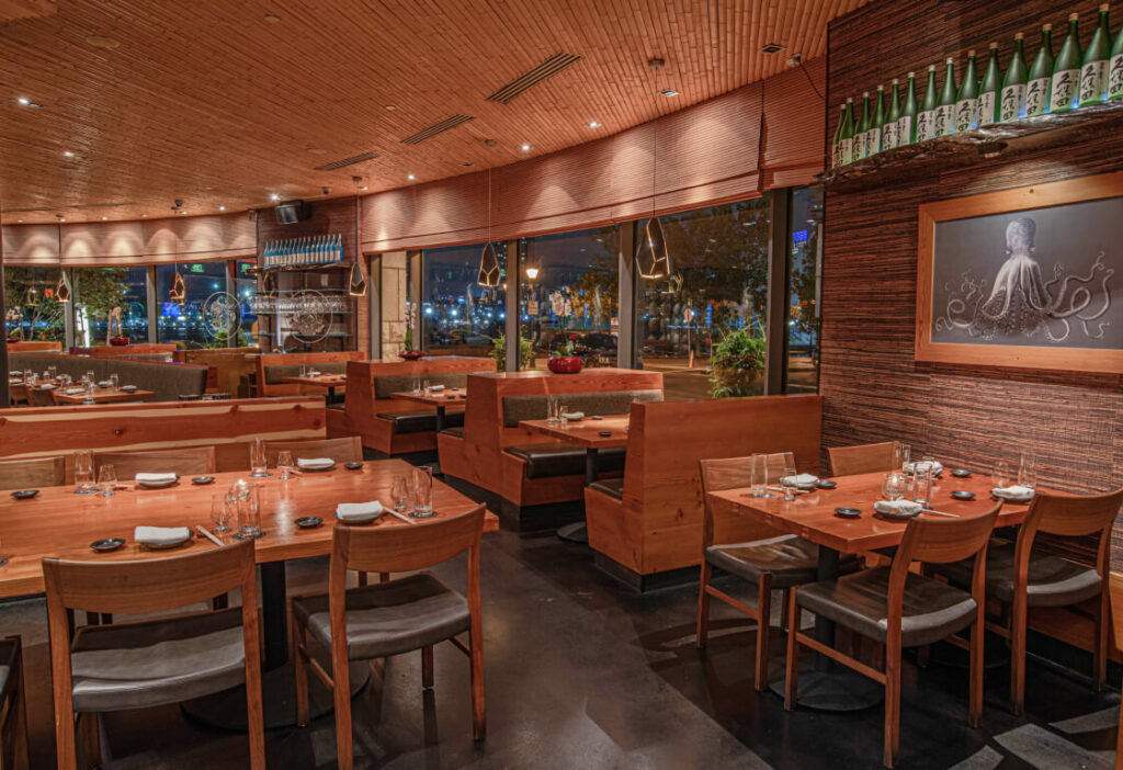 A view of a modern restaurant interior featuring wooden tables and chairs, booths, neatly set tables, a large window, and a wall adorned with bottles and a picture of an octopus.