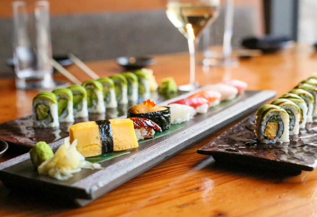 A variety of sushi rolls and nigiri arranged on two rectangular plates, accompanied by a glass of white wine, soy sauce, wasabi, and pickled ginger, on a wooden table.