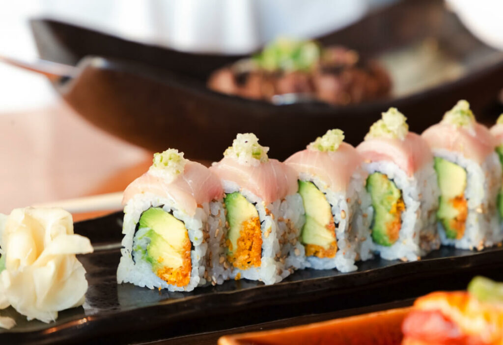 A plate of sushi rolls with avocado, rice, and fish, topped with a small amount of wasabi. Pickled ginger is placed on the side. A blurred dish is in the background.