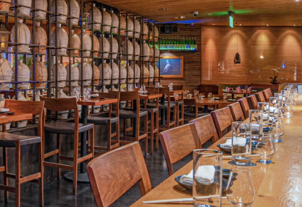 A restaurant interior with wooden tables and chairs. The left wall is lined with barrels, and there are place settings of glasses, dishes, and napkins on the tables. A painting hangs on the far wall.