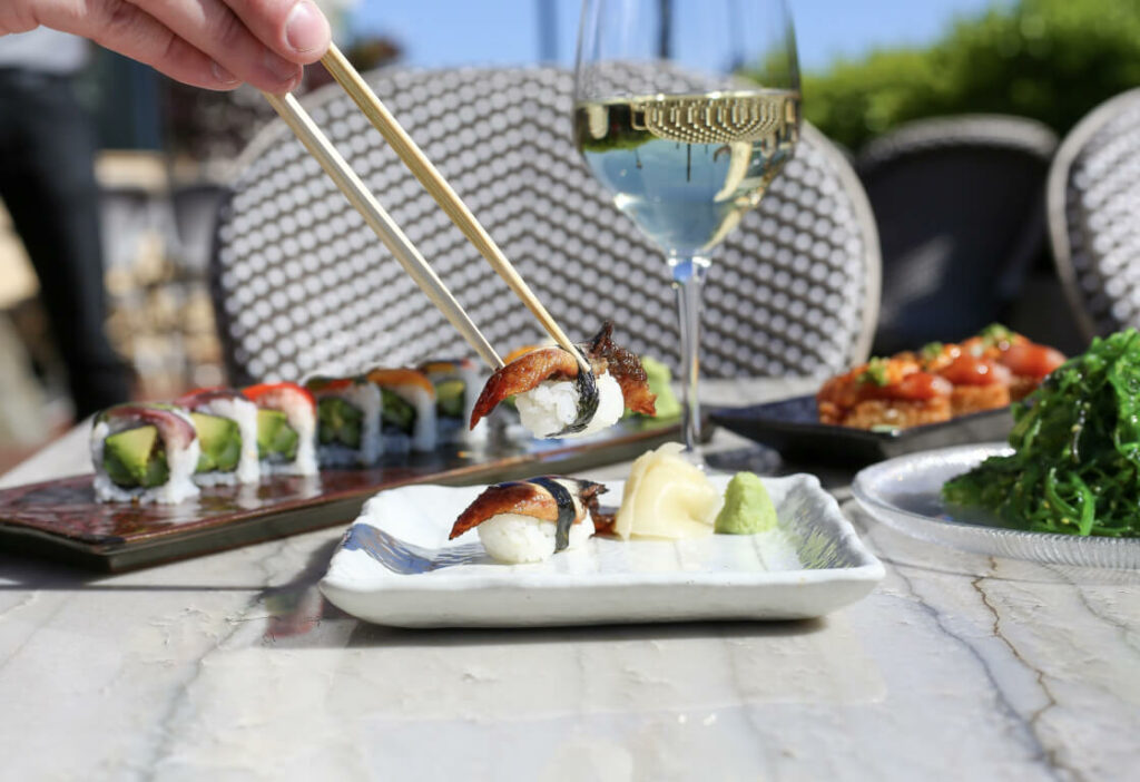 A hand holding sushi with chopsticks over a plate on a table with various sushi dishes and a glass of white wine in an outdoor setting.