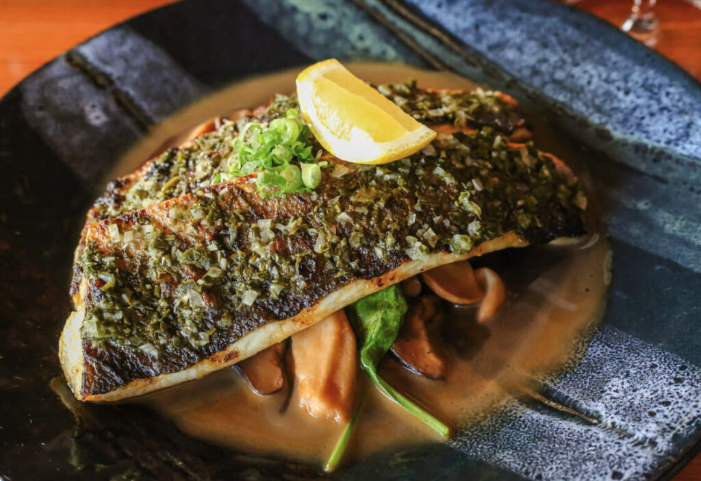 Grilled fish fillet topped with herbs and a lemon wedge, served over sautéed vegetables and sauce, presented on a dark ceramic plate.