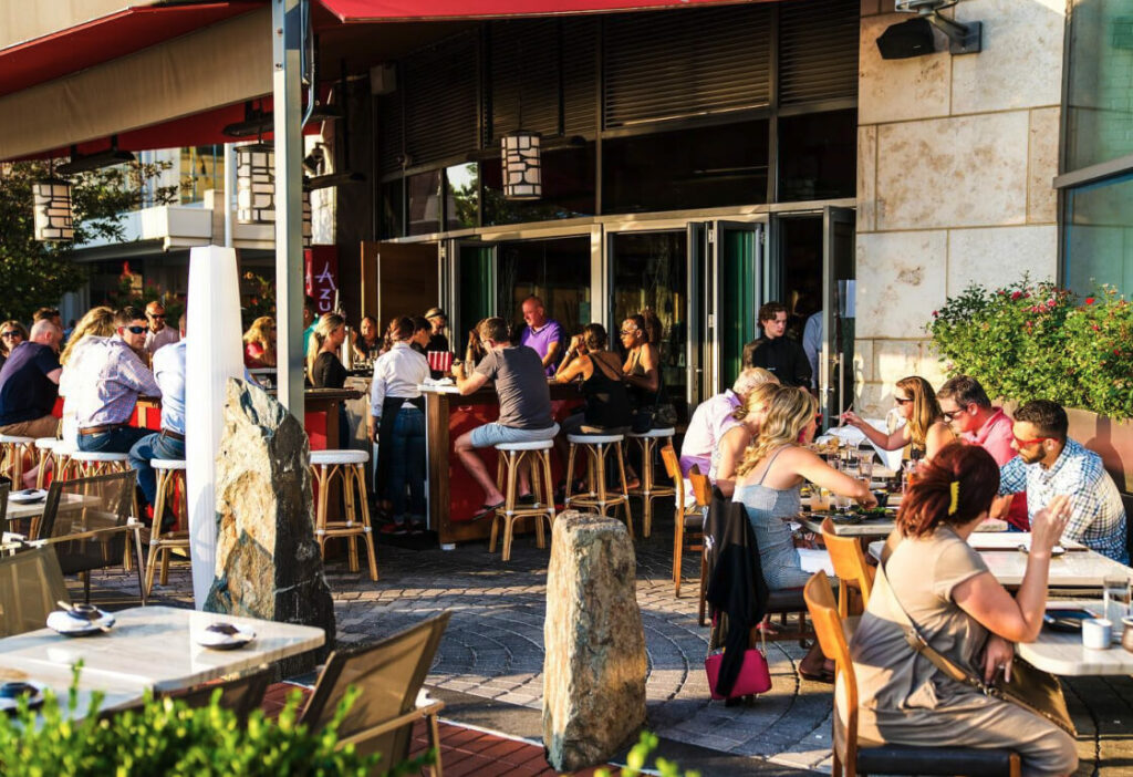 People are seated and enjoying food and drinks at an outdoor restaurant on a sunny day. Tables and bar seating are visible, with patrons socializing and engaging in conversations.