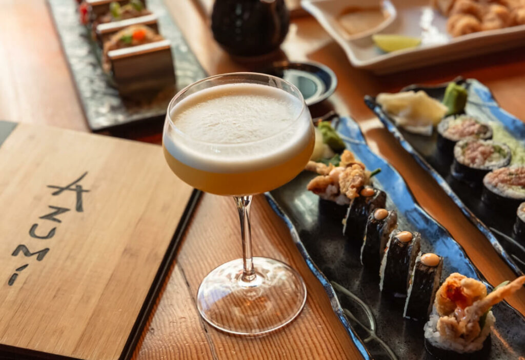 A cocktail in a coupe glass next to a wooden menu and three plates of sushi, displayed on a wooden table.