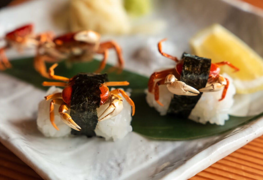 Two pieces of sushi topped with small whole crabs and wrapped in seaweed placed on a rectangular plate with a lemon wedge and garnishes in the background.