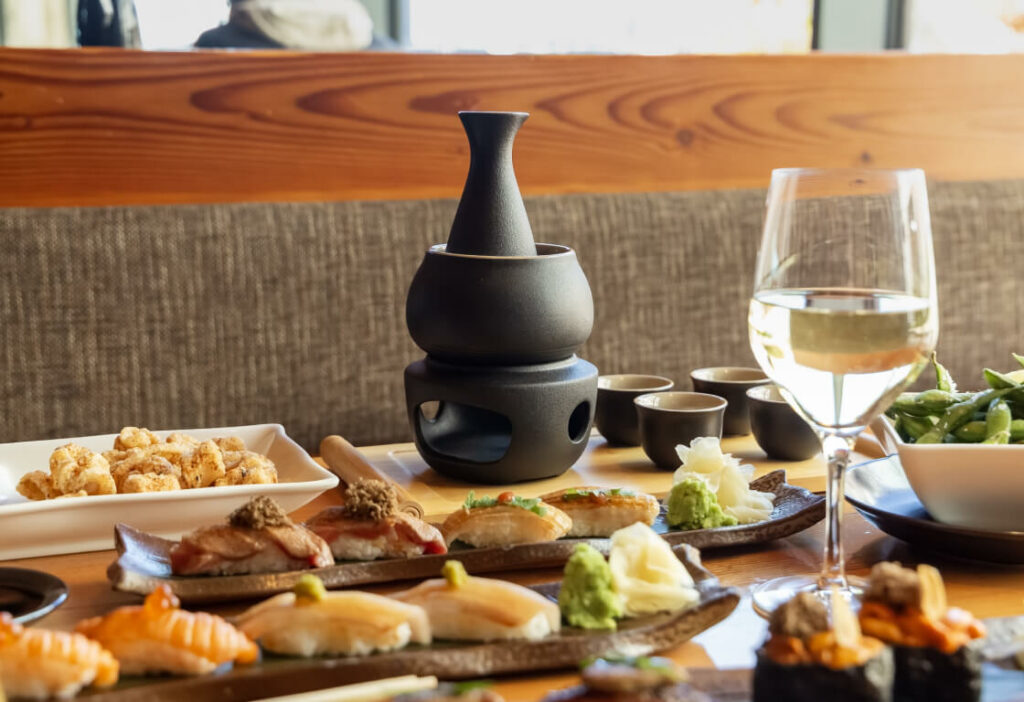 A variety of sushi rolls, a glass of white wine, a sake set, and bowls of edamame and fried appetizers on a wooden table at a restaurant.