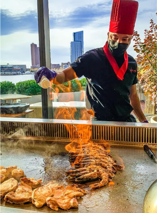 A chef wearing a red hat and mask cooks shrimp and chicken on a grill, with flames rising from the hot surface. City buildings and waterfront are visible in the background.