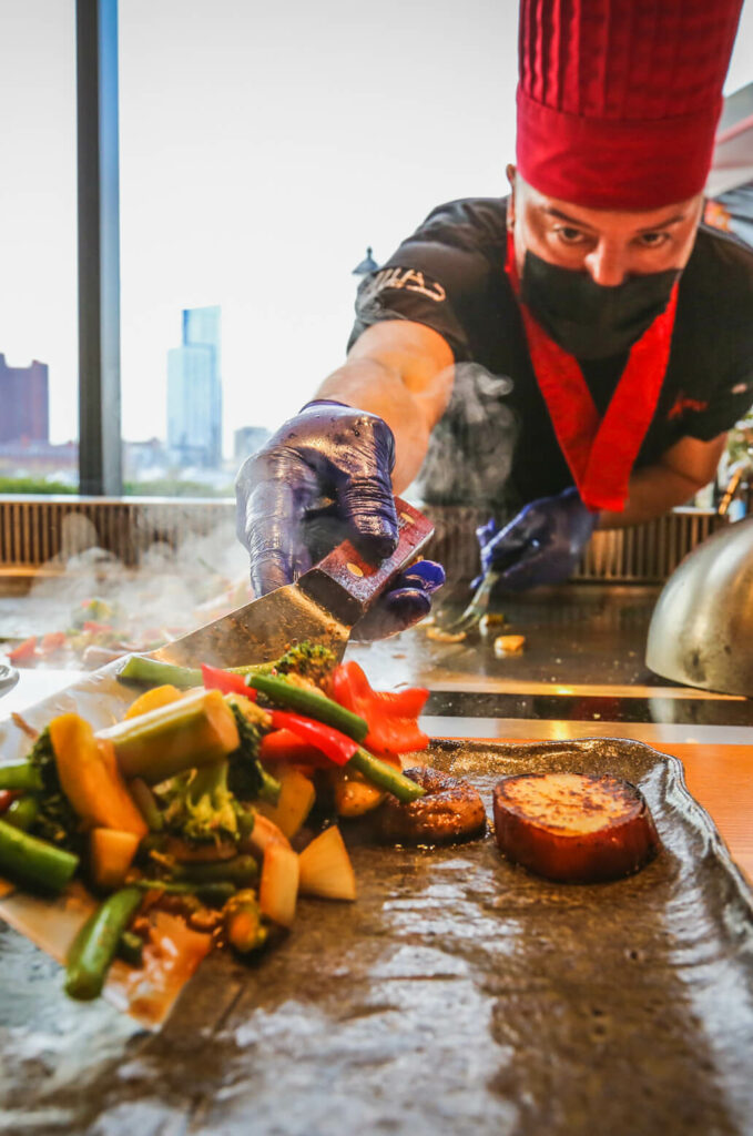 A chef wearing a red hat and black mask prepares grilled vegetables on a griddle, using a spatula to arrange a mix of broccoli, peppers, and other vegetables.