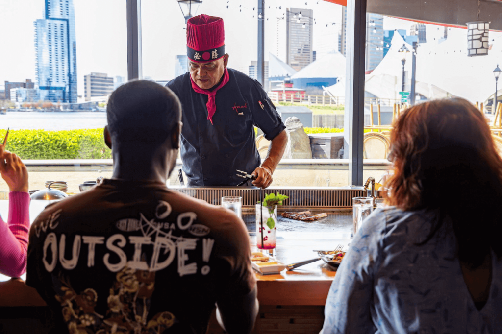 A chef in a red hat and black uniform prepares food on a grill at a restaurant, while three patrons seated at the counter watch. City buildings and waterfront are visible through the window.
