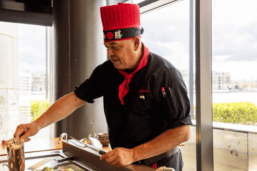 Chef in a red hat and black uniform prepares food on a teppanyaki grill in front of a window with a cityscape view.