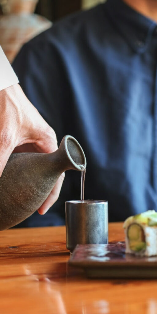 A hand is pouring liquid from a ceramic pitcher into a small cup. Part of a sushi roll is visible on a plate in the foreground, and a person in a dark shirt is blurred in the background.