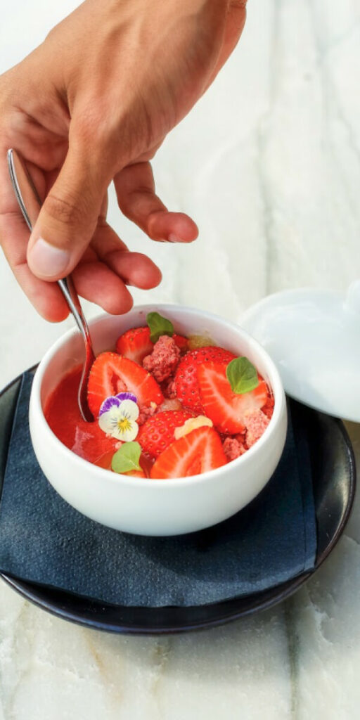 A hand holding a spoon over a white bowl filled with strawberries, small green leaves, and a small flower garnish. The bowl rests on a dark napkin on a plate.