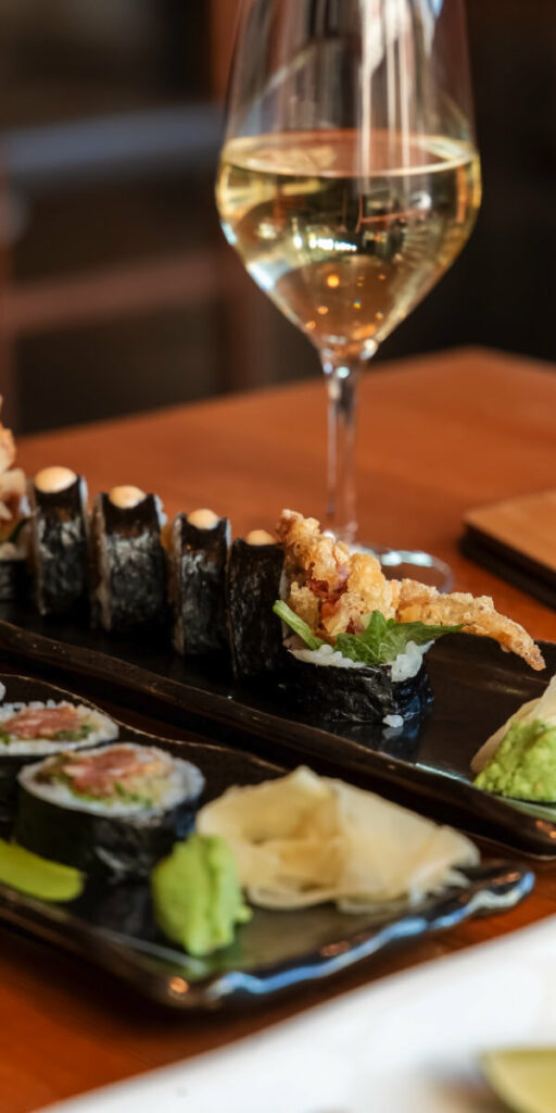 A close-up of a sushi platter featuring various rolls and nigiri, garnished with wasabi and pickled ginger. A glass of white wine is in the background on a wooden table.