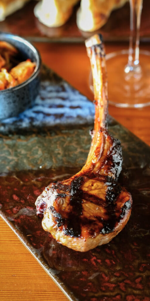 A cooked lamb chop with grill marks is served on a rectangular plate with a side dish in a small bowl.