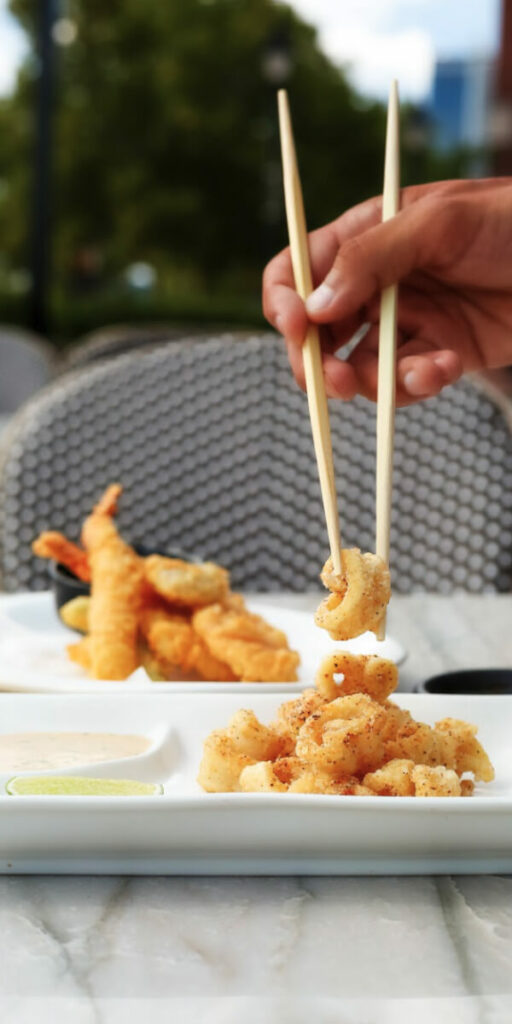 A hand using chopsticks to pick a piece of fried food from a plate, with another dish containing tempura in the background.