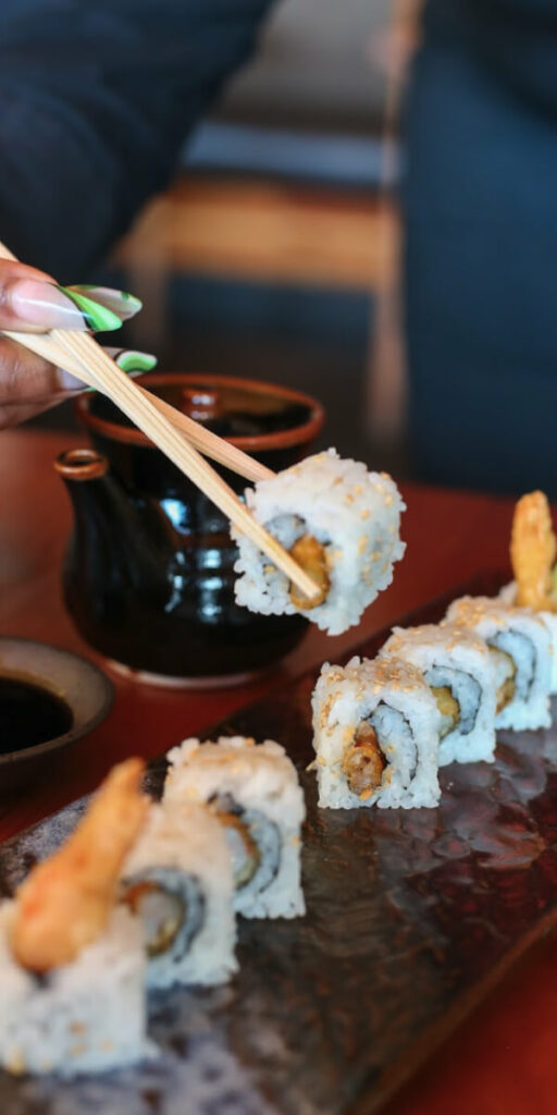 A hand holding chopsticks picks up a piece of sushi roll from a plate containing several pieces. A small sauce dish and a dark teapot are in the background.