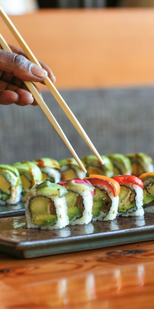 A hand holding chopsticks is picking up a piece of sushi from a plate with several colorful sushi rolls on a wooden table.