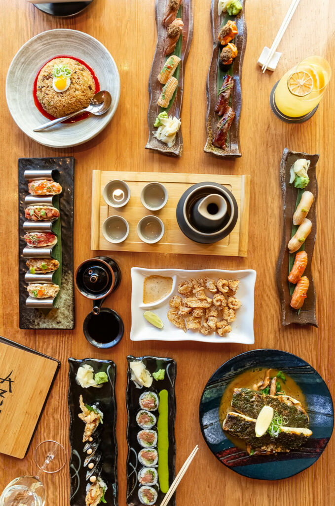 A variety of Japanese dishes including sushi rolls, sashimi, takoyaki, and a hot beverage set arranged on a wooden table. Drinks include an orange slice garnished beverage.