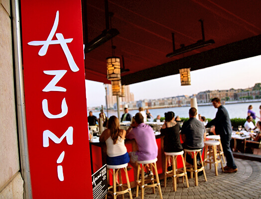 People sit at a bar counter in an outdoor restaurant with red decor and a waterfront view. A red panel with the word 