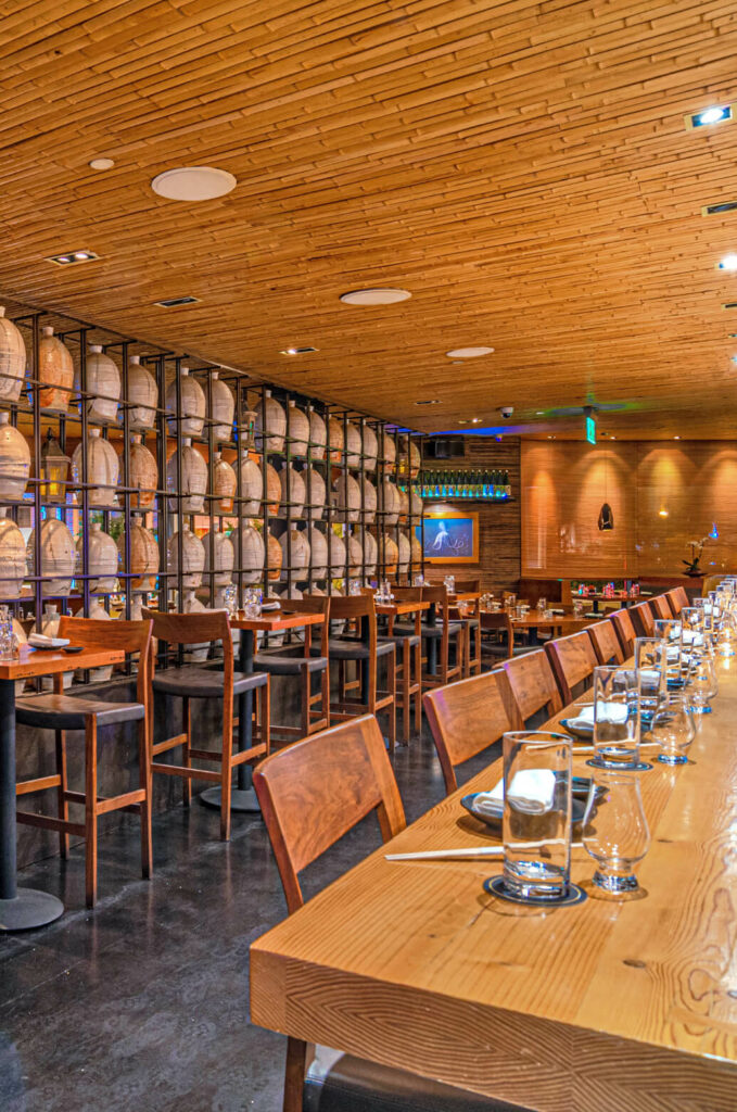 A restaurant interior with wooden tables and chairs, glassware arranged on tables, a wall of barrels, and warm, ambient lighting under a wooden ceiling. An illuminated blue sign is visible in the background.