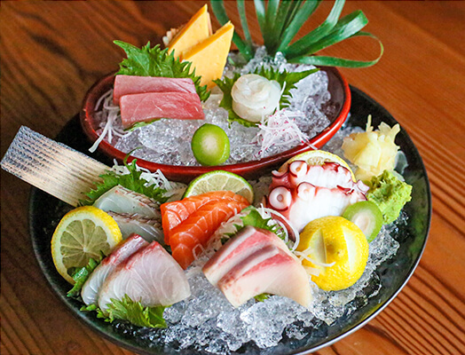 A plate of sashimi on ice with various types of fish, lemon slices, shiso leaves, wasabi, and garnish, arranged on a wooden table.