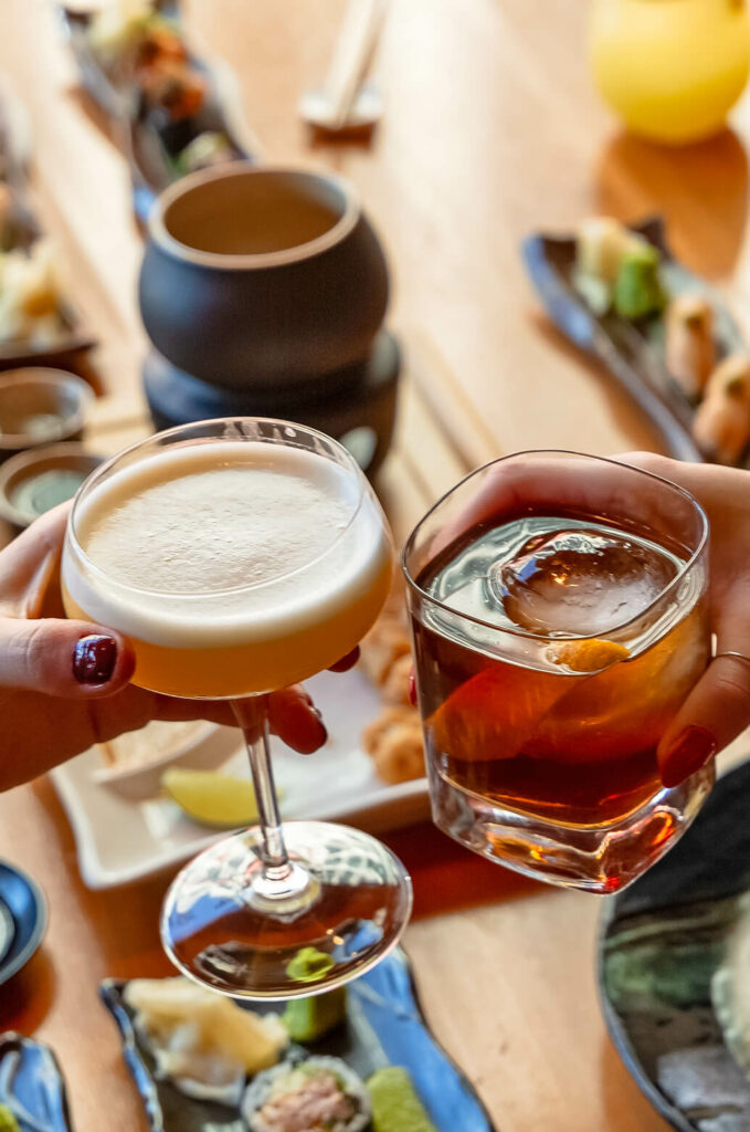 Two people holding cocktails in a restaurant, one with a frothy white drink in a coupe glass and the other with a dark beverage in a rocks glass with an ice cube. Food dishes are visible in the background.