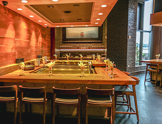 Cozy restaurant bar with wooden chairs and a wooden counter, light fixtures on the ceiling, and neatly set tables with flowers. A wide window shows an outdoor view.