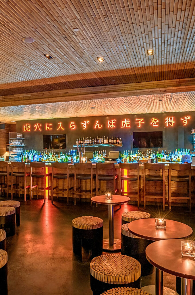 A modern bar with a well-lit counter, bottles displayed on shelves, and neon Japanese characters on the wall. Wooden stools and tables are arranged in the dimly lit seating area.