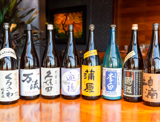 An assortment of eight sake bottles with various labels are placed in a row on a wooden surface, set against a blurred indoor background with a painting on the wall.