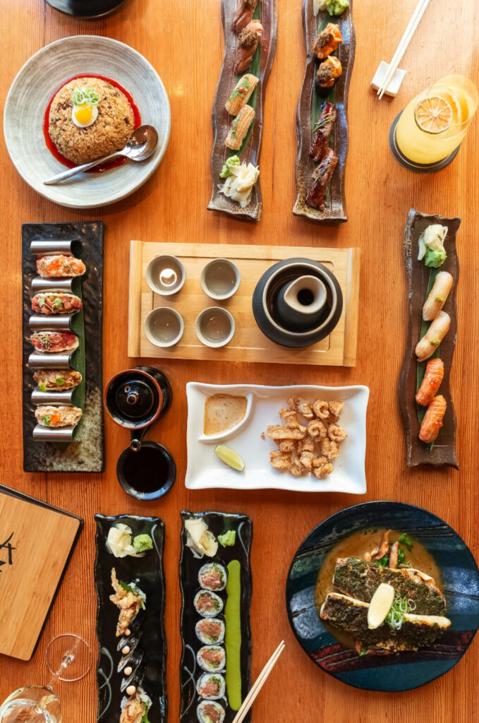 An overhead view of a variety of sushi dishes, rolls, fried rice, grilled seafood, and a beverage arranged on a wooden table with chopsticks.