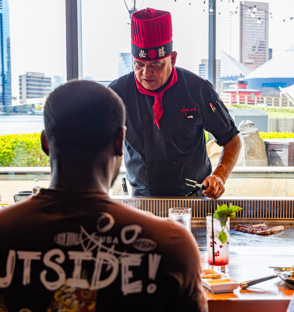 A chef in a red hat and black uniform prepares food on a teppanyaki grill, with a customer seated in front wearing a brown shirt that reads 