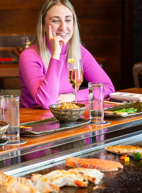 Woman in a pink sweater smiling and sitting at a restaurant table with grilled seafood, a bowl of food, glass of water, and a glass of sparkling wine in front of her.