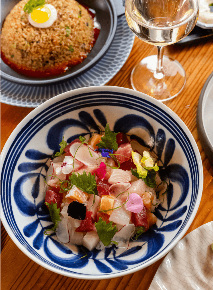 A blue-and-white bowl filled with a colorful poke bowl sits on a wooden table next to a glass of white wine and a plate of fried rice topped with a quail egg.