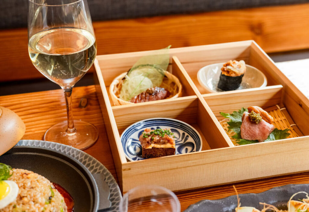 A wooden box with four compartments, each holding different Japanese dishes, is placed next to a glass of white wine on a wooden table. Other assorted dishes are partially visible around it.