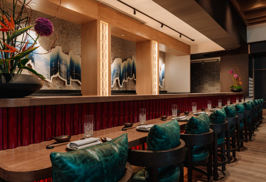 A modern restaurant interior with a long wooden table, teal upholstered chairs, neatly set tableware, and contemporary wall art with floral arrangements visible.