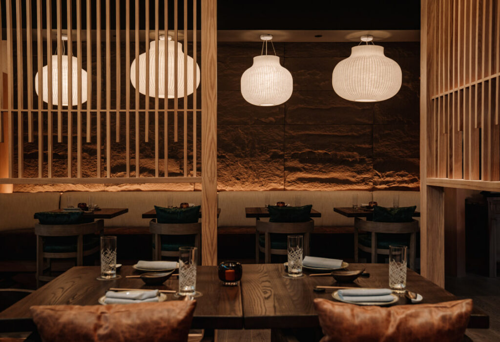 A dimly lit restaurant interior with wooden dividers, modern pendant lights, and set tables with glassware and plates.