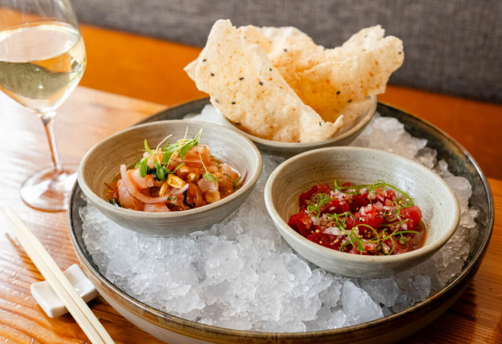 A plate with bowls of poke, fresh vegetables, and crispy rice chips on ice, served with a glass of white wine and chopsticks on the side.