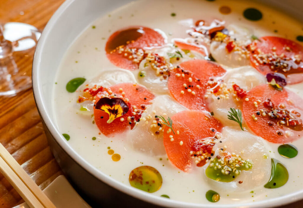 A bowl of gourmet soup with colorful garnishes including white broth, red slices, green droplets, edible flowers, and sprinkled seeds, served with wooden chopsticks on the side.
