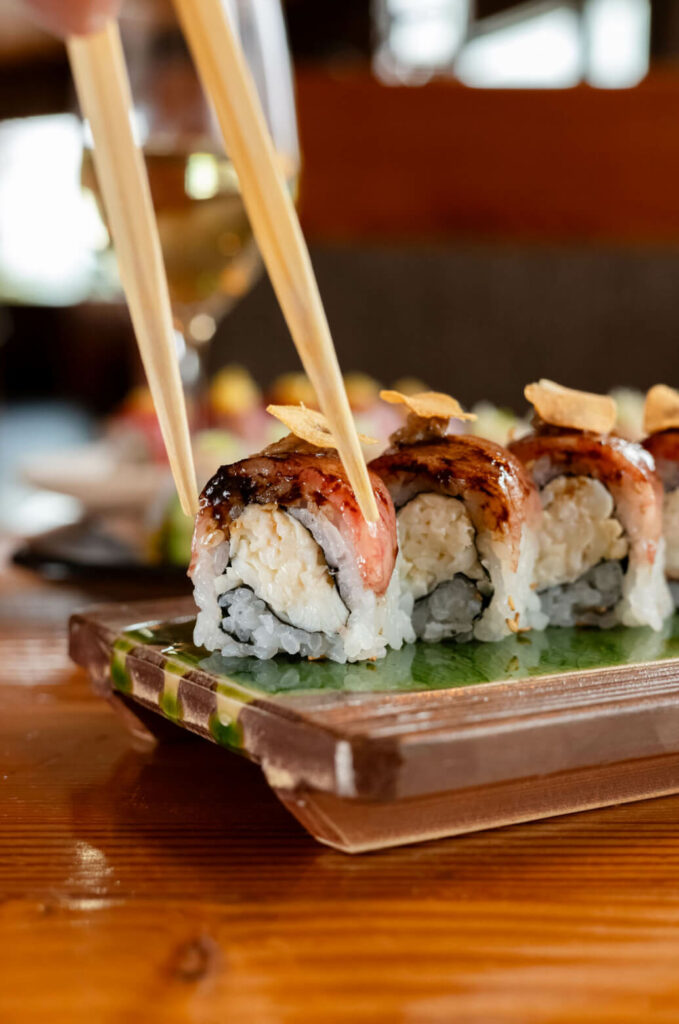 Close-up of a pair of chopsticks holding a piece of sushi from a wooden tray. The sushi has visible layers of rice, fish, and a topping, and a glass of white wine is in the blurred background.