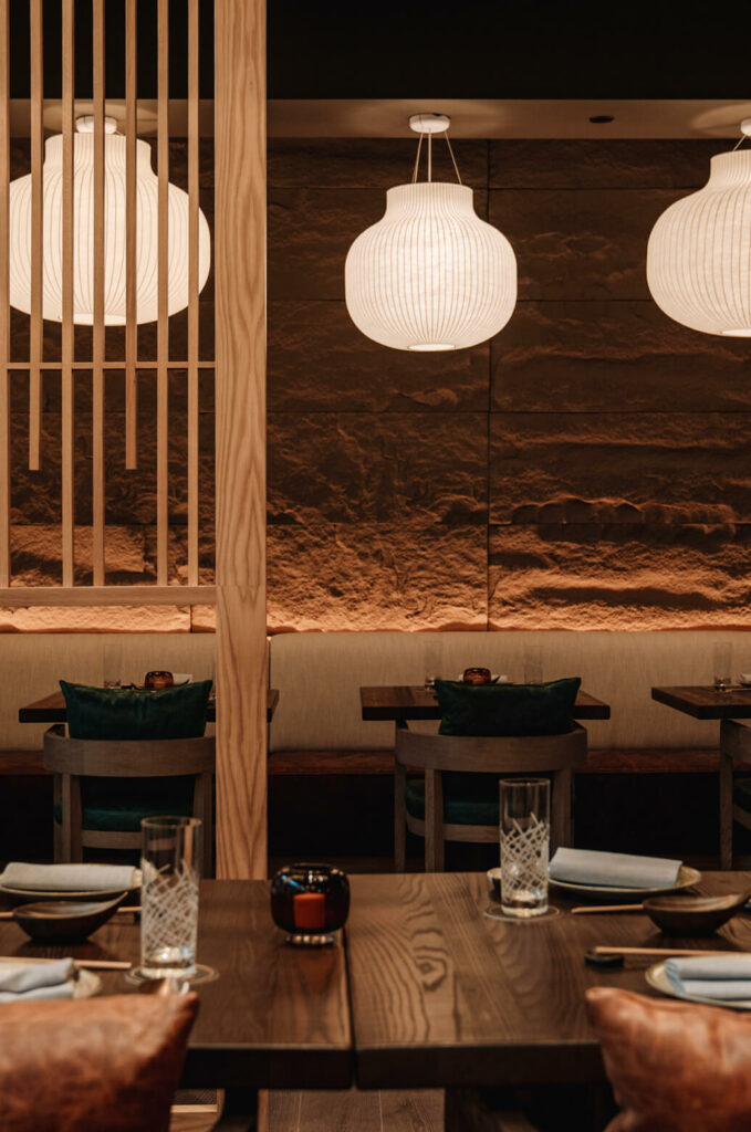 A dimly lit restaurant with wooden tables and chairs, soft glowing pendant lights, and textured wall panels, creating an intimate dining atmosphere.