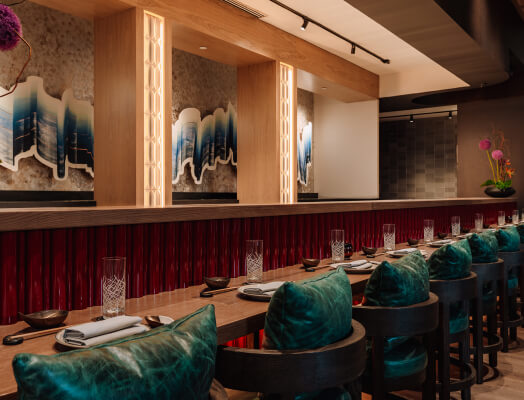 An upscale restaurant interior featuring a long wooden counter with green cushioned stools, neatly arranged cutlery, and decorative wall art illuminated by warm lighting.