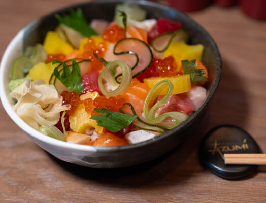 A bowl of colorful sushi ingredients including fish, rice, vegetables, and roe, garnished with ginger. Chopsticks and a round black cap with the word 