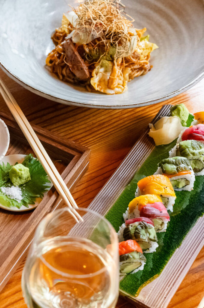A bowl of noodles with vegetables and meat, a wooden tray with assorted sushi rolls, a glass of white wine, and chopsticks placed on a wooden table.