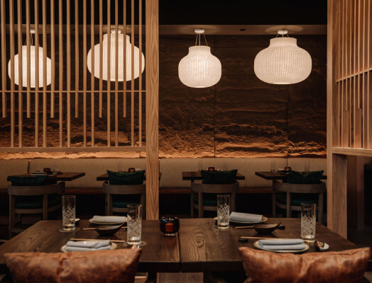 A dimly lit restaurant features wooden partitions, hanging paper lanterns, and neatly set tables with glassware and napkins, creating a cozy and intimate dining ambiance.