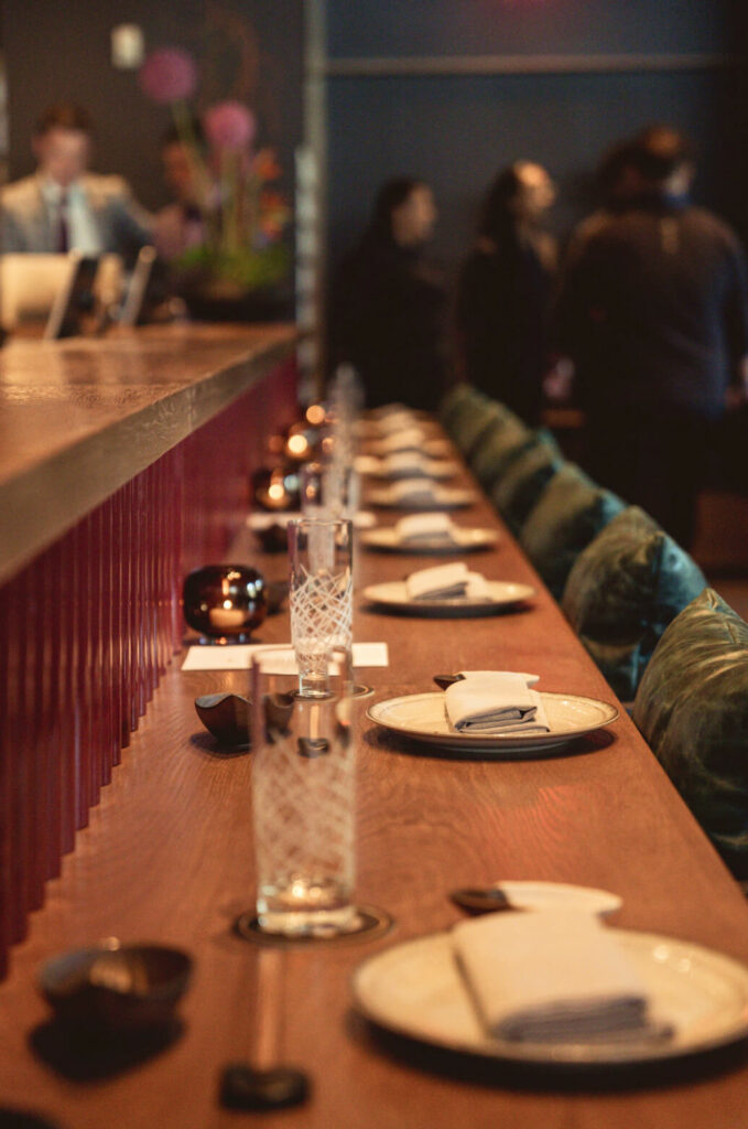 A restaurant bar with a wooden counter is set with plates, glassware, napkins, and chopsticks. Several people stand conversing in the background.