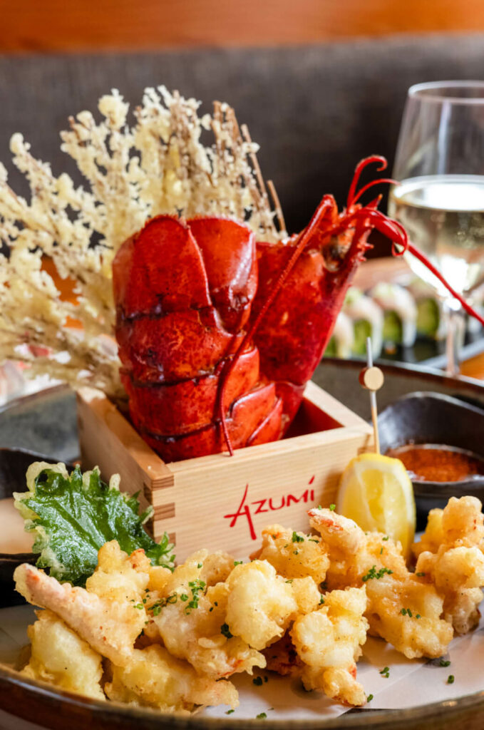 A plate of fried seafood garnished with greenery next to a wooden box containing a cooked lobster tail, with a glass of white wine in the background.
