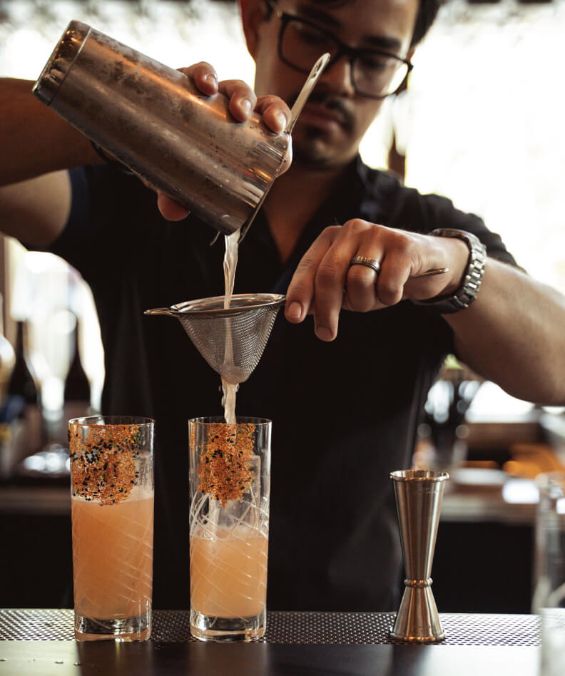 Bartender pouring a mixed drink through a strainer into two glasses, each with a garnish, on a bar counter. Measures and equipment are visible.