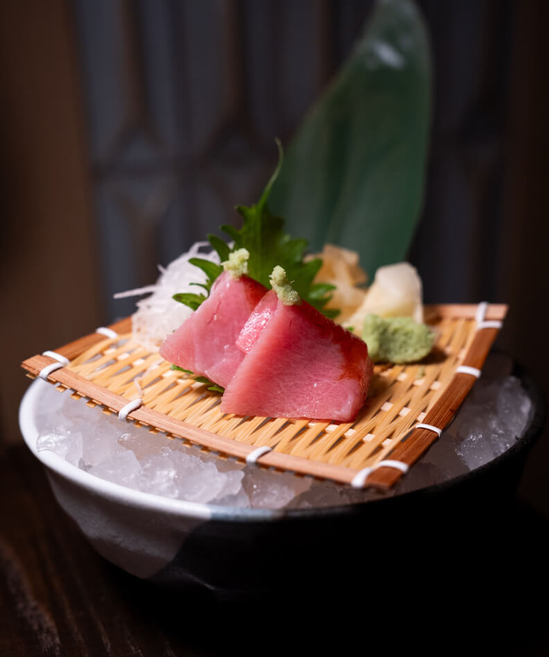 A plate of sashimi, featuring two pieces of tuna garnished with wasabi, and thinly sliced radish, served on a bamboo mat over a bowl of ice.
