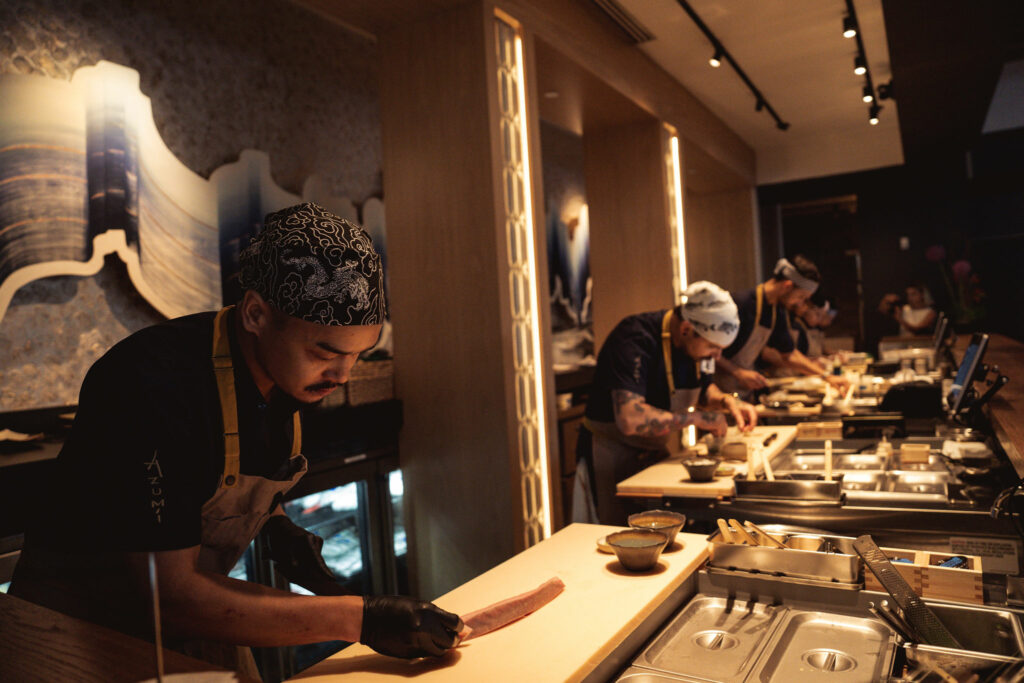 Chefs in a modern kitchen are preparing dishes behind a counter. One chef in the foreground focuses on slicing a piece of fish. The background shows other chefs engaged in their tasks.