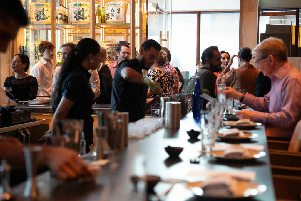 A busy bar scene with bartenders preparing drinks and patrons sitting and standing at the counter, engaging in conversation. The background showcases illuminated shelves of bottles.